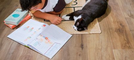 A boy reading on a rug on the floor with his pet cat keeping him company.