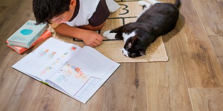 A boy reading on a rug on the floor with his pet cat keeping him company.