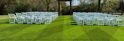 Artificial grass event lawn with white seating for outdoor ceremony Outdoor wedding ceremony setup with white chairs on manicured artificial turf lawn
