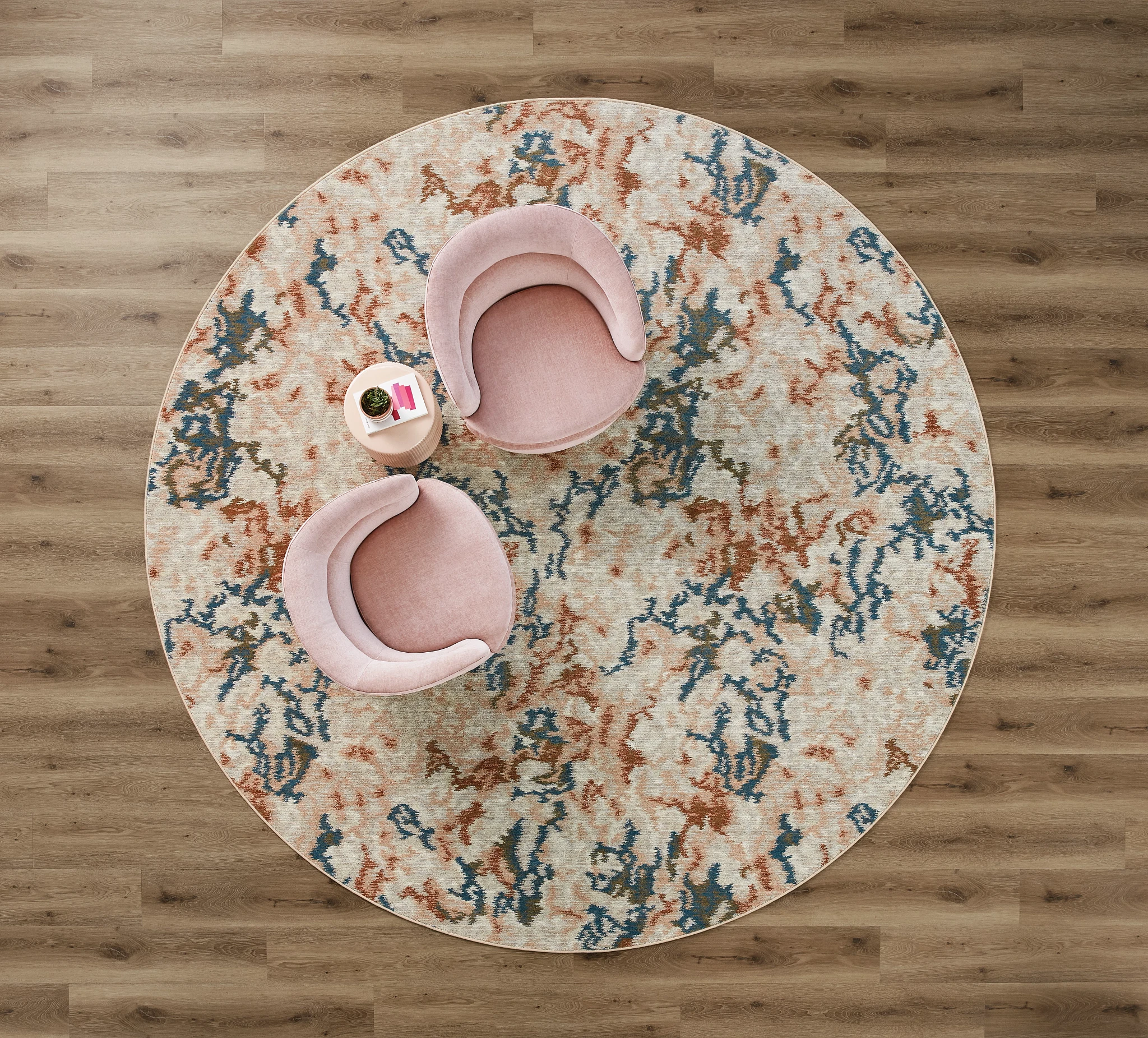 Overhead view of a modern room with light wood flooring and a round patterned rug featuring pink, blue, and beige colors, accompanied by two pink chairs and a small round table