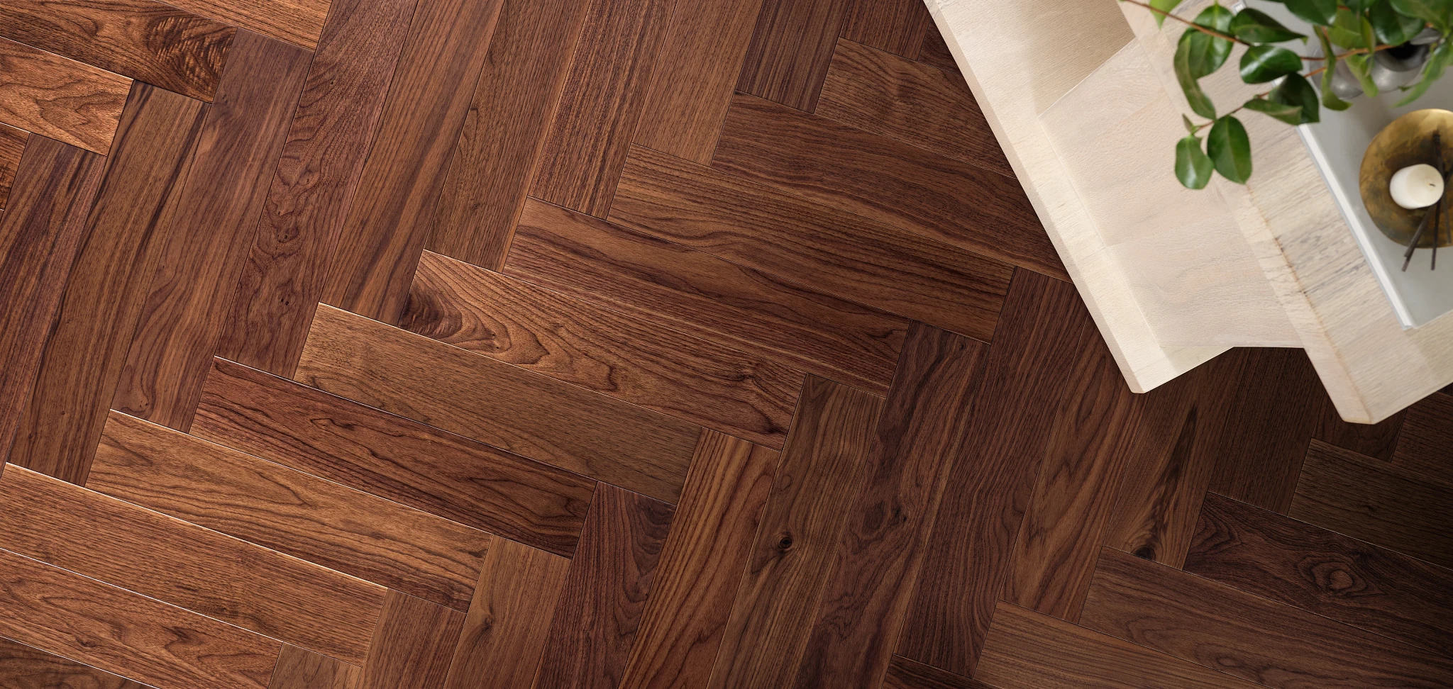 Herringbone pattern hardwood flooring in rich brown tones with a light wood table and green plant